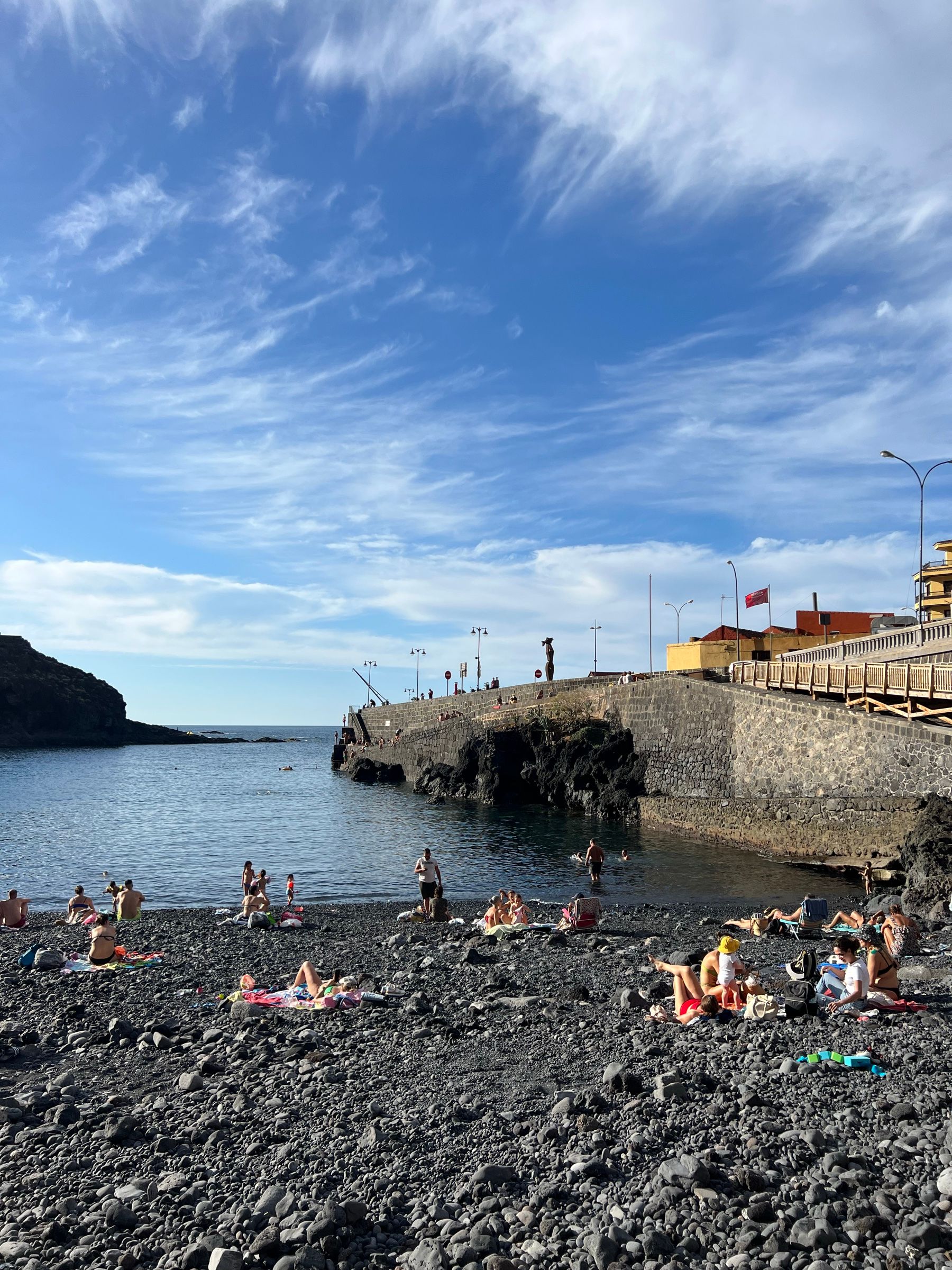 La Playa del Muelle de Garachico llena de gente sobre los callaos negros, con el muelle viejo al fondo.