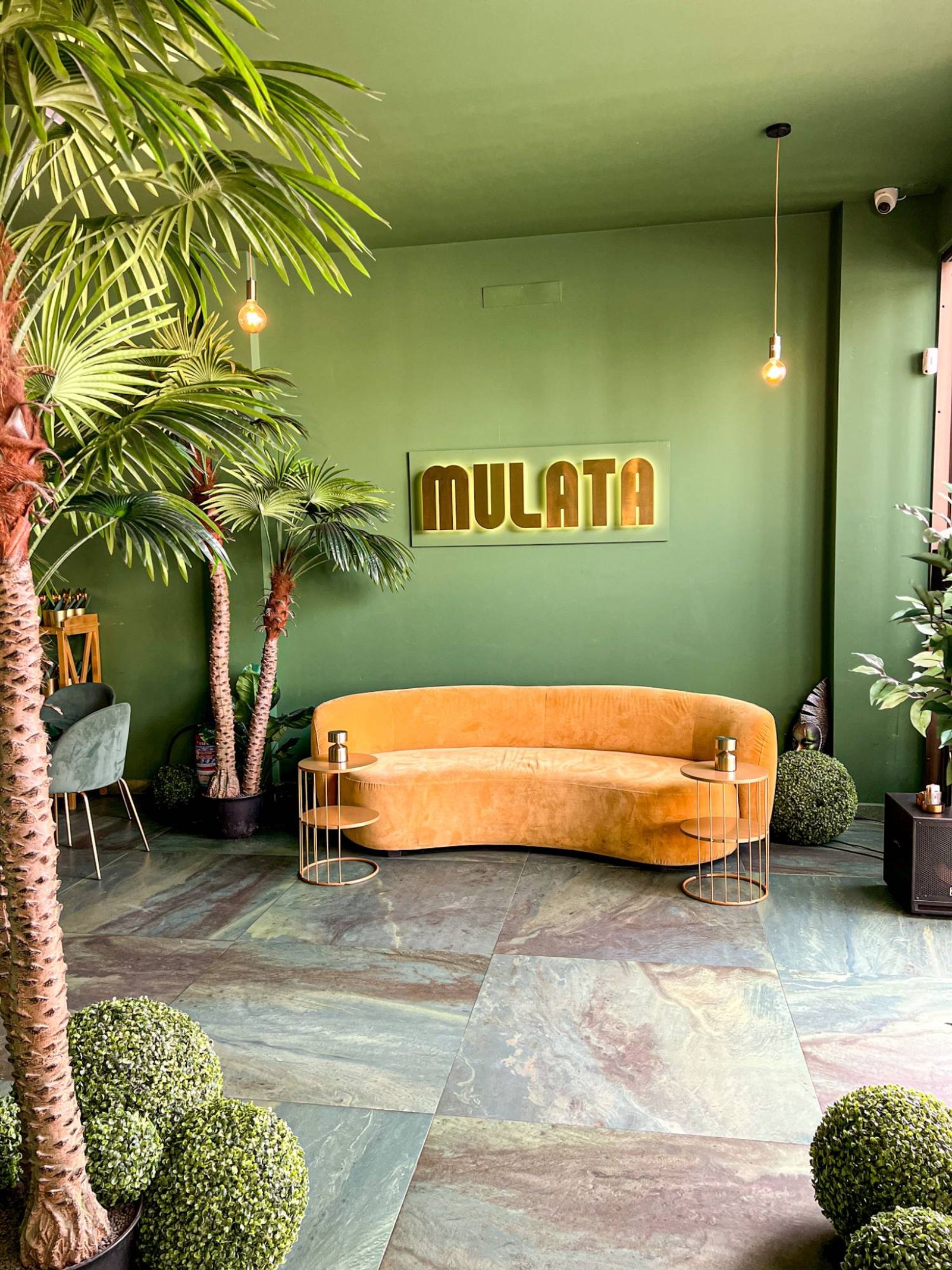 A curved yellow sofa under MULATA's gold sign on the green wall, with palms either side, small gold tables and a marble floor.