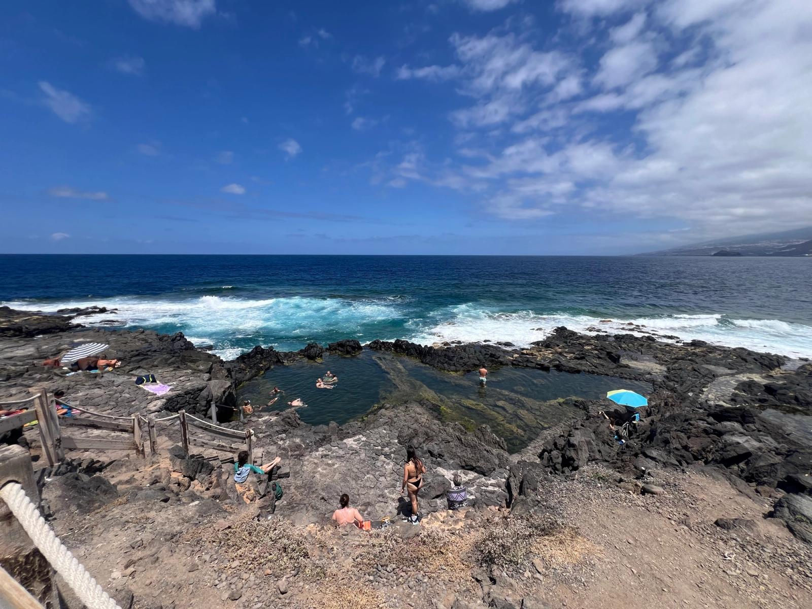 Vista panorámica del Charco de los Chochos (Charco Los Chochos), la piscina natural de Los Silos, en Tenerife: la poza de roca volcánica con gente bañándose, la escalera de acceso y las olas rompiendo detrás.
