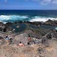 Vista panorámica del Charco de los Chochos (Charco Los Chochos), la piscina natural de Los Silos, en Tenerife: la poza de roca volcánica con gente bañándose, la escalera de acceso y las olas rompiendo detrás.