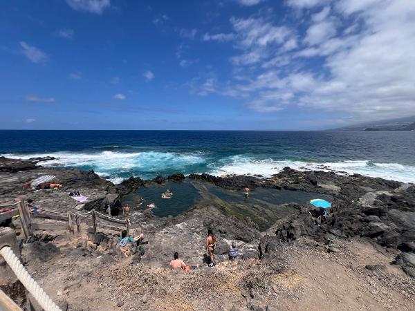 Vista panorámica del Charco de los Chochos (Charco Los Chochos), la piscina natural de Los Silos, en Tenerife: la poza de roca volcánica con gente bañándose, la escalera de acceso y las olas rompiendo detrás.
