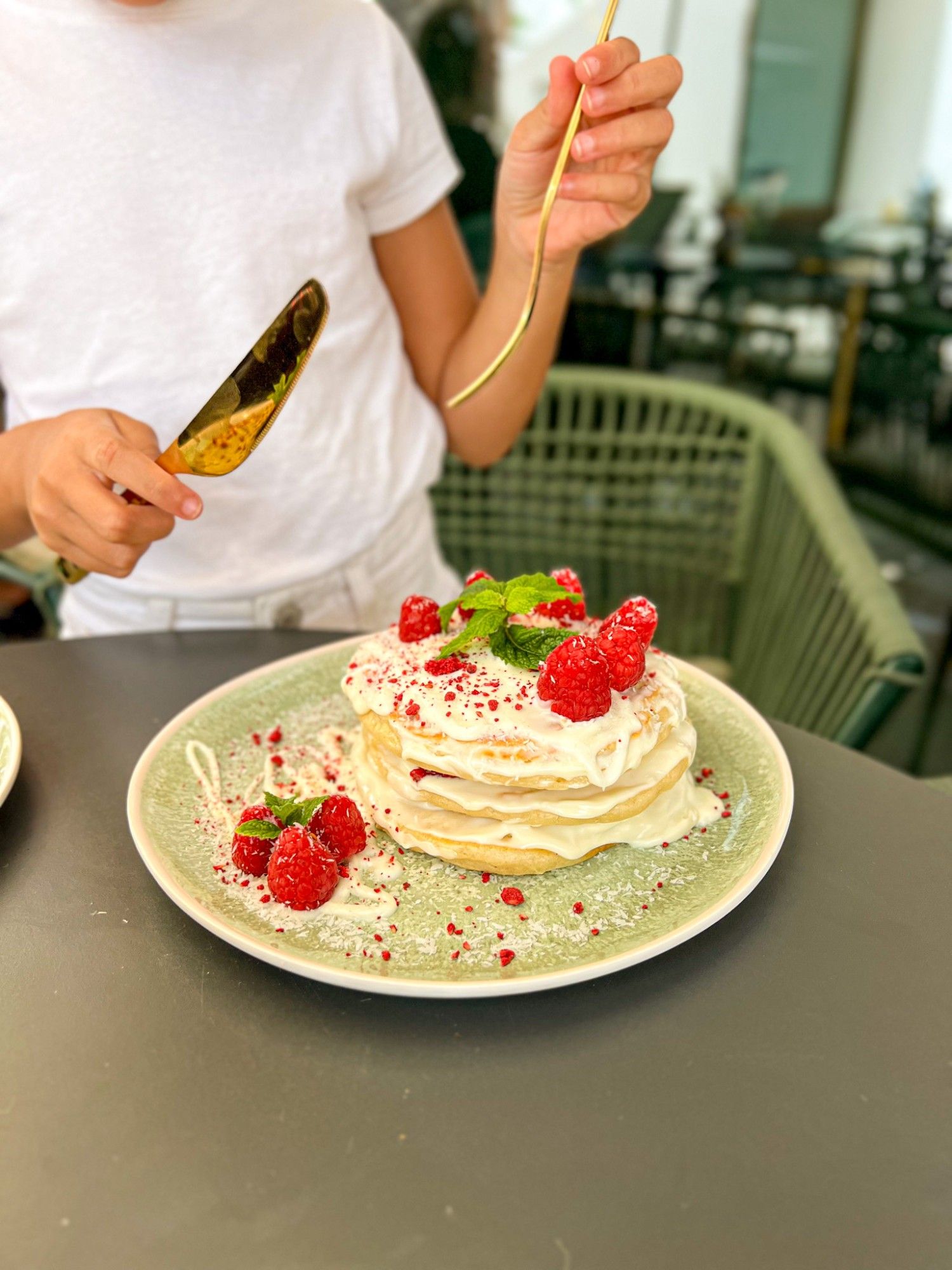 White chocolate pancakes with cream, fresh raspberries, grated coconut and mint on a green plate, with a girl about to dig in with a gold spoon.