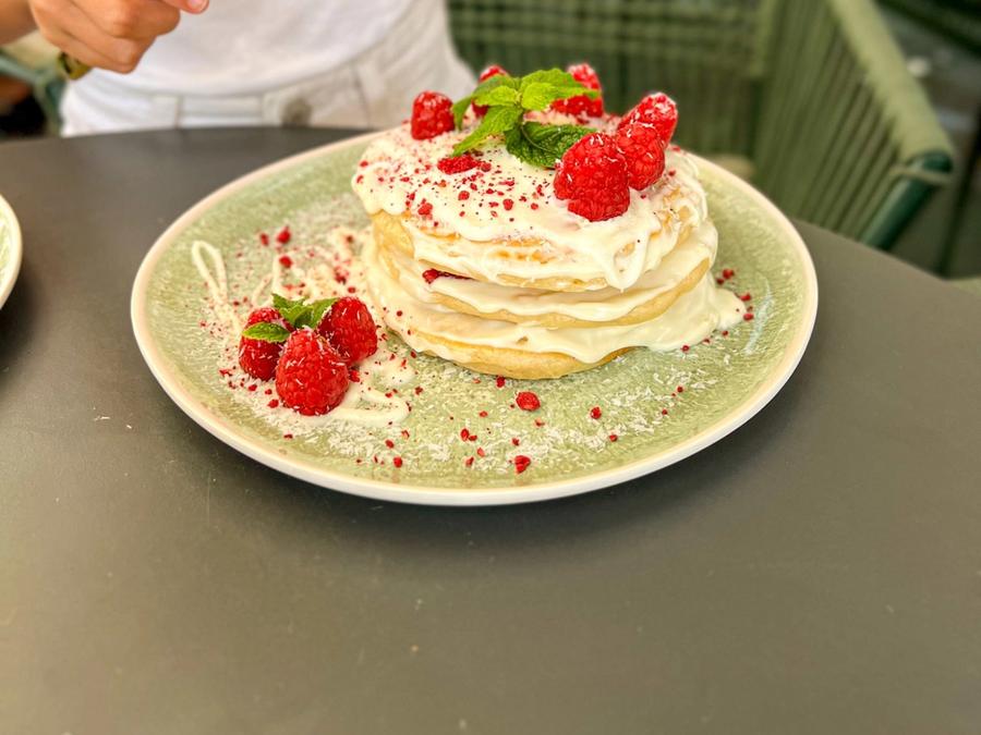 White chocolate pancakes with cream, fresh raspberries, grated coconut and mint on a green plate, with a girl about to dig in with a gold spoon.