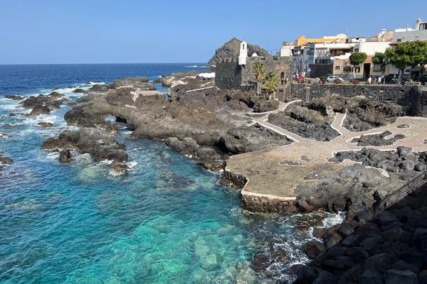 El agua turquesa de las piscinas naturales de El Caletón, en Garachico, entre la roca volcánica y con las casas del pueblo al fondo.