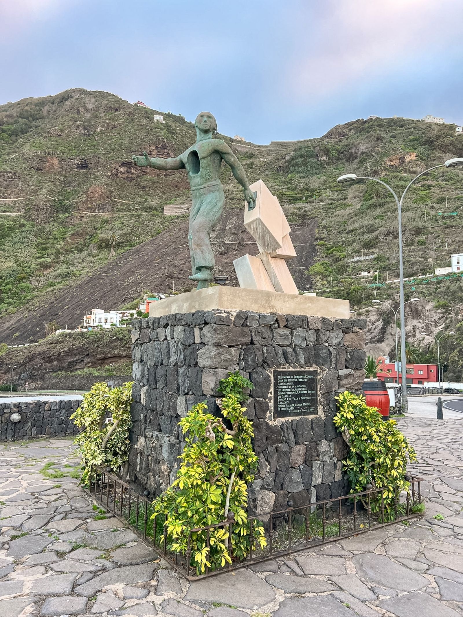 Monumento al Emigrante Canario en Garachico: figura de bronce con el brazo extendido sobre una base de piedra volcánica, con la montaña detrás.