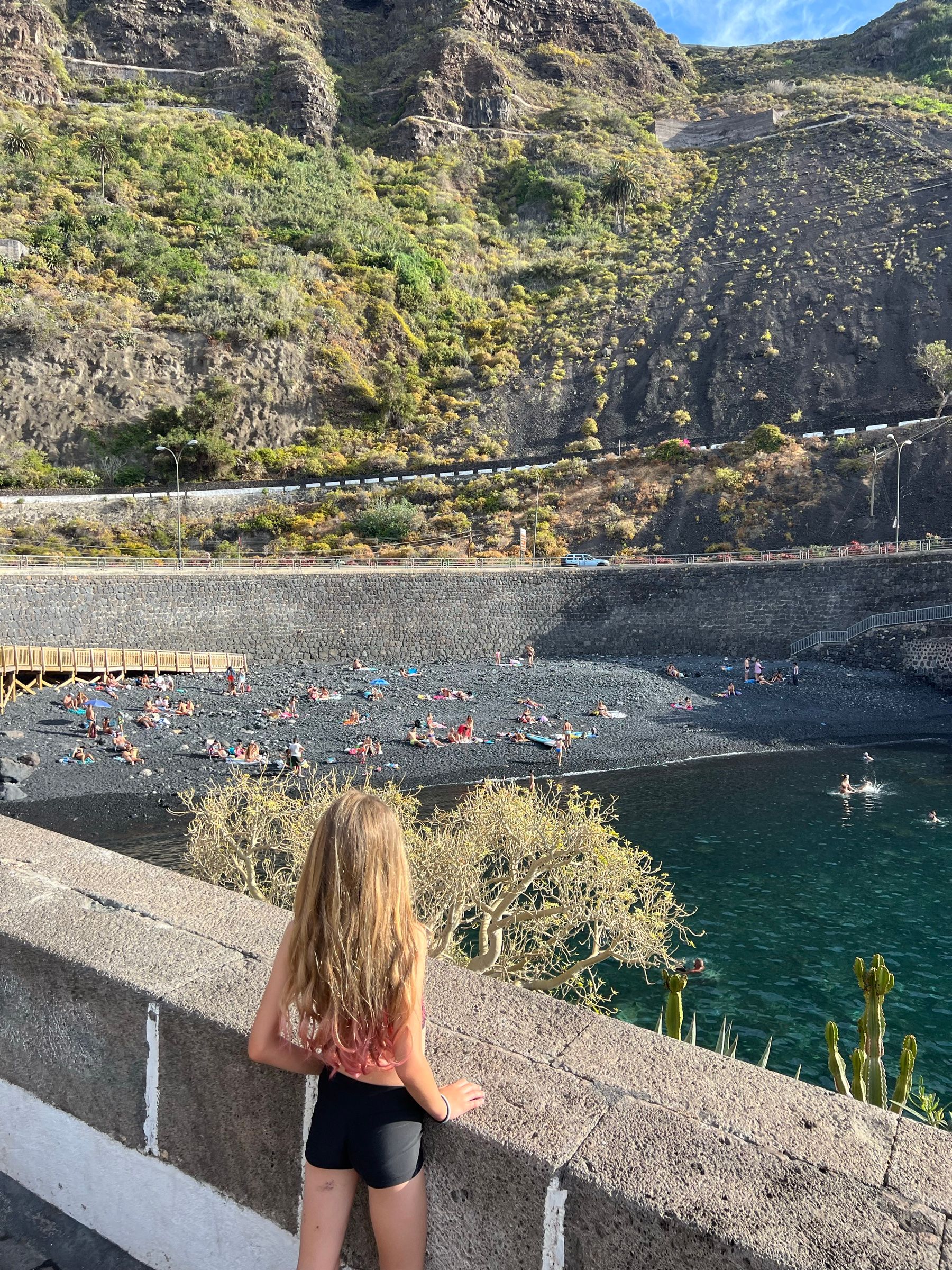 Una niña mira desde el muro la Playa del Muelle de Garachico, con la ladera verde y el agua clara abajo.