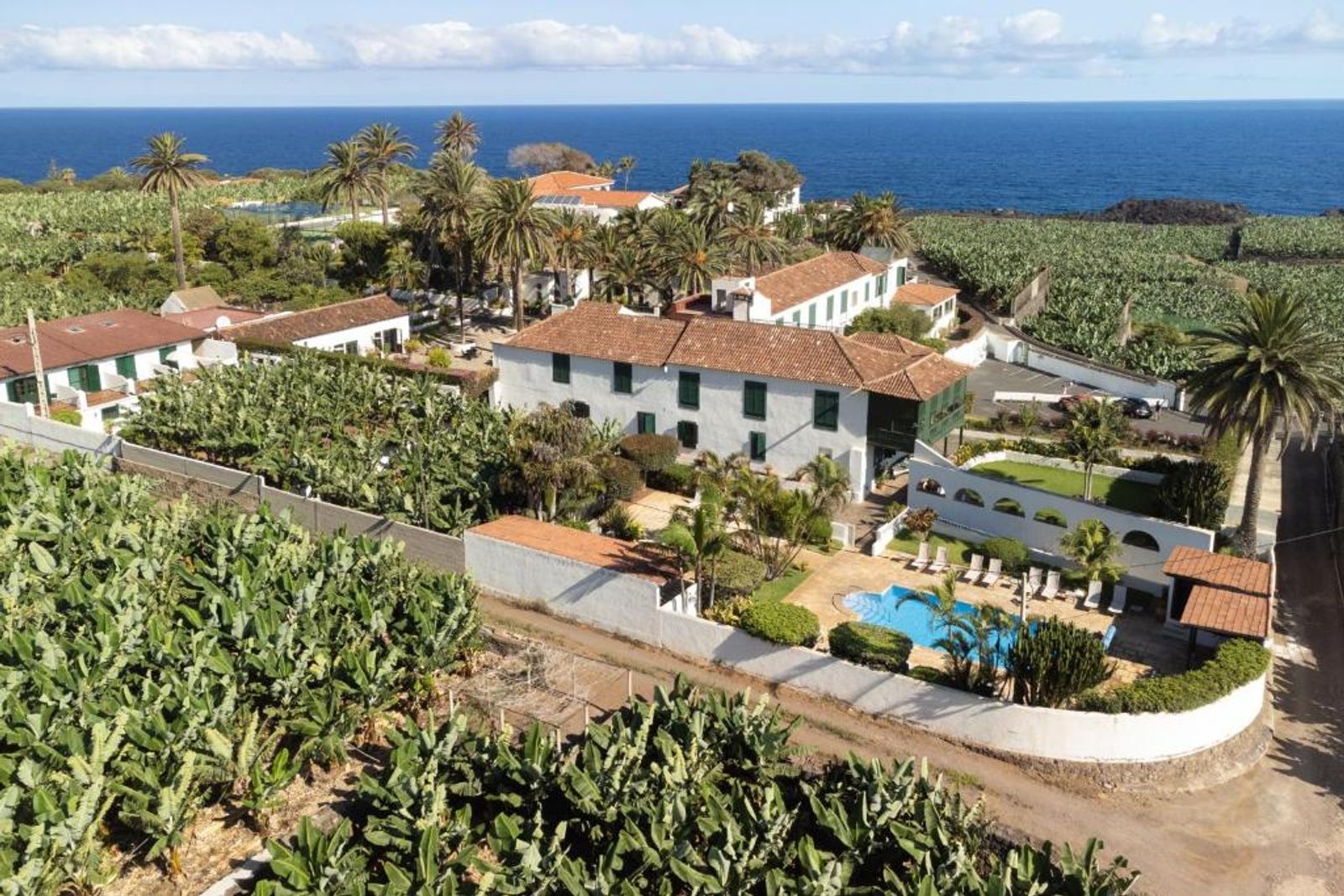 Vista aérea del Hotel Rural El Patio, en Garachico: la casona blanca de tejas rojas rodeada de plataneras, con la piscina, las palmeras y el Atlántico al fondo.