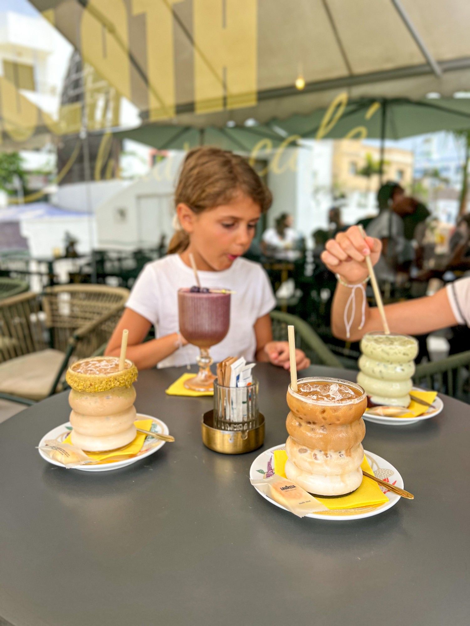 A table on MULATA's terrace with a purple smoothie, an iced pistachio latte, an iced caramel latte and a matcha latte in ribbed glasses, with a girl drinking behind.