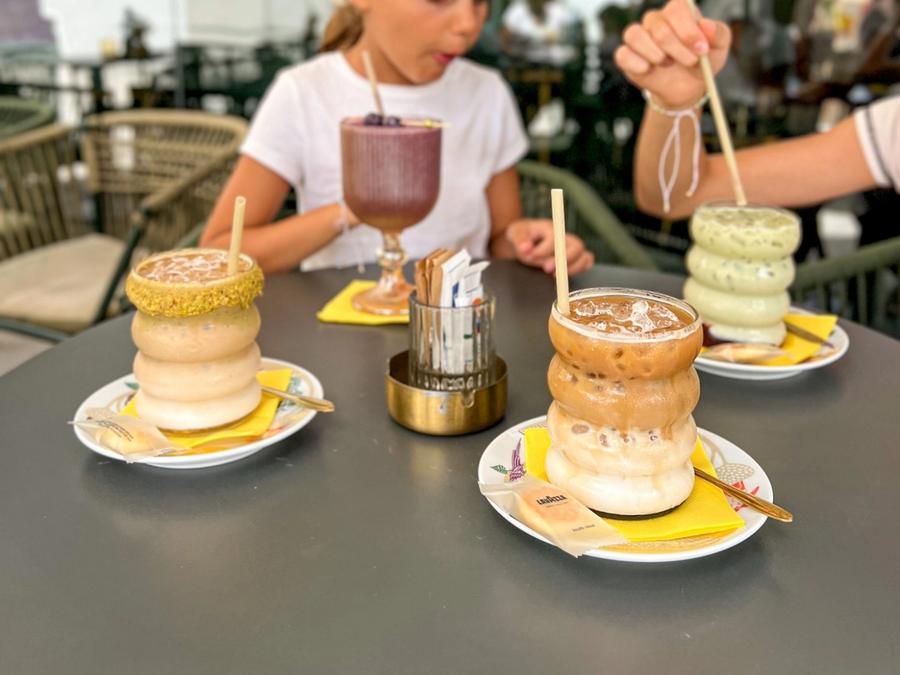 A table on MULATA's terrace with a purple smoothie, an iced pistachio latte, an iced caramel latte and a matcha latte in ribbed glasses, with a girl drinking behind.