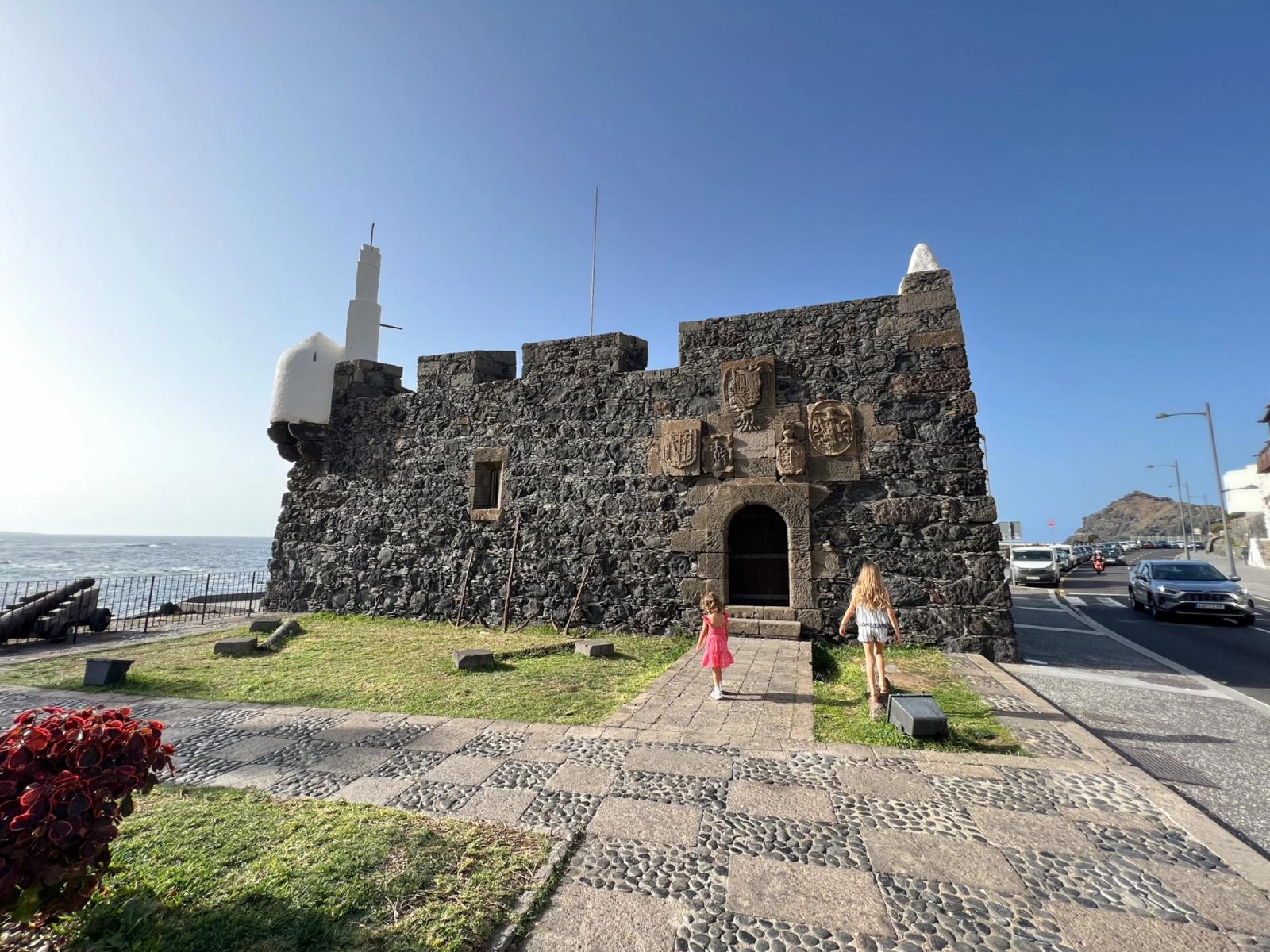 El Castillo de San Miguel de Garachico visto desde el paseo, con los cañones sobre la hierba, el mar a un lado y dos niñas subiendo hacia la puerta.
