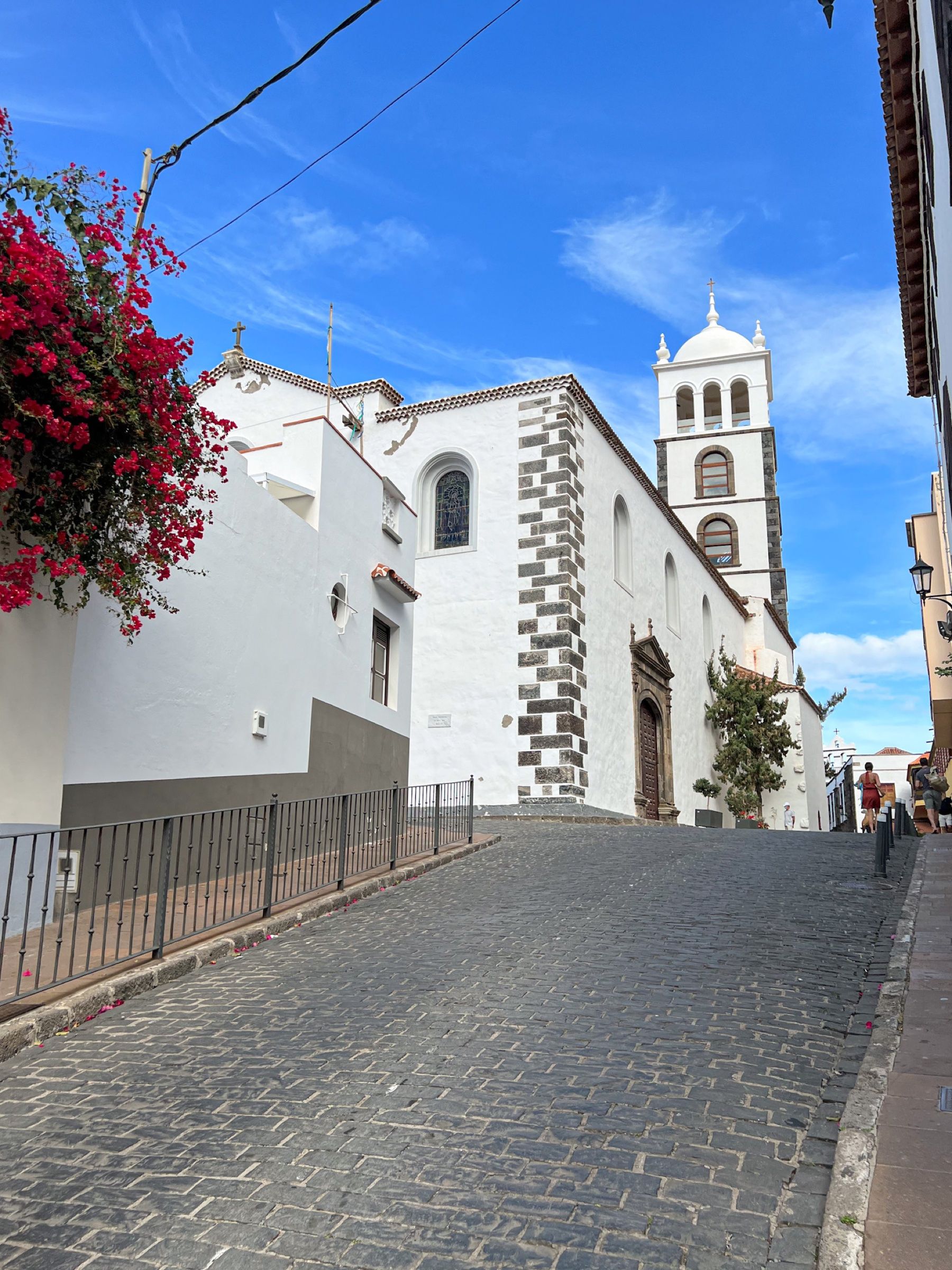 Calle empedrada de Garachico subiendo junto a la iglesia de Santa Ana, con su torre blanca, las esquinas de piedra volcánica y una buganvilla en flor.