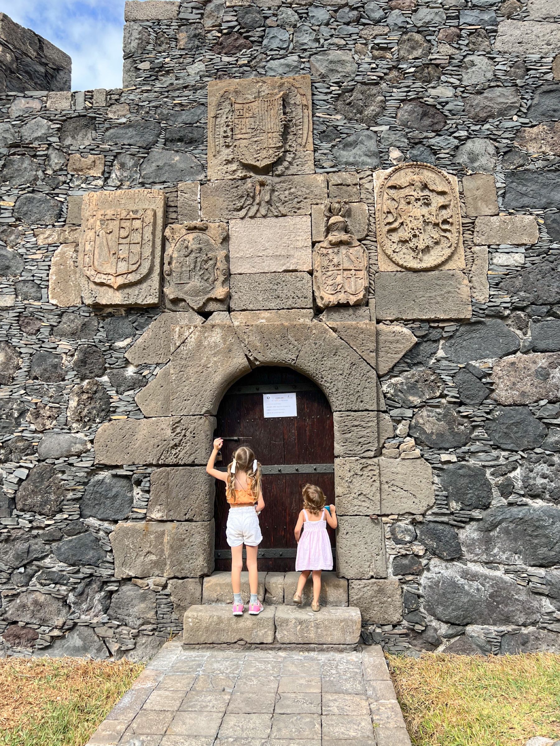 Dos niñas asomadas a la puerta de madera del Castillo de San Miguel de Garachico, bajo los escudos de piedra tallados en el muro.