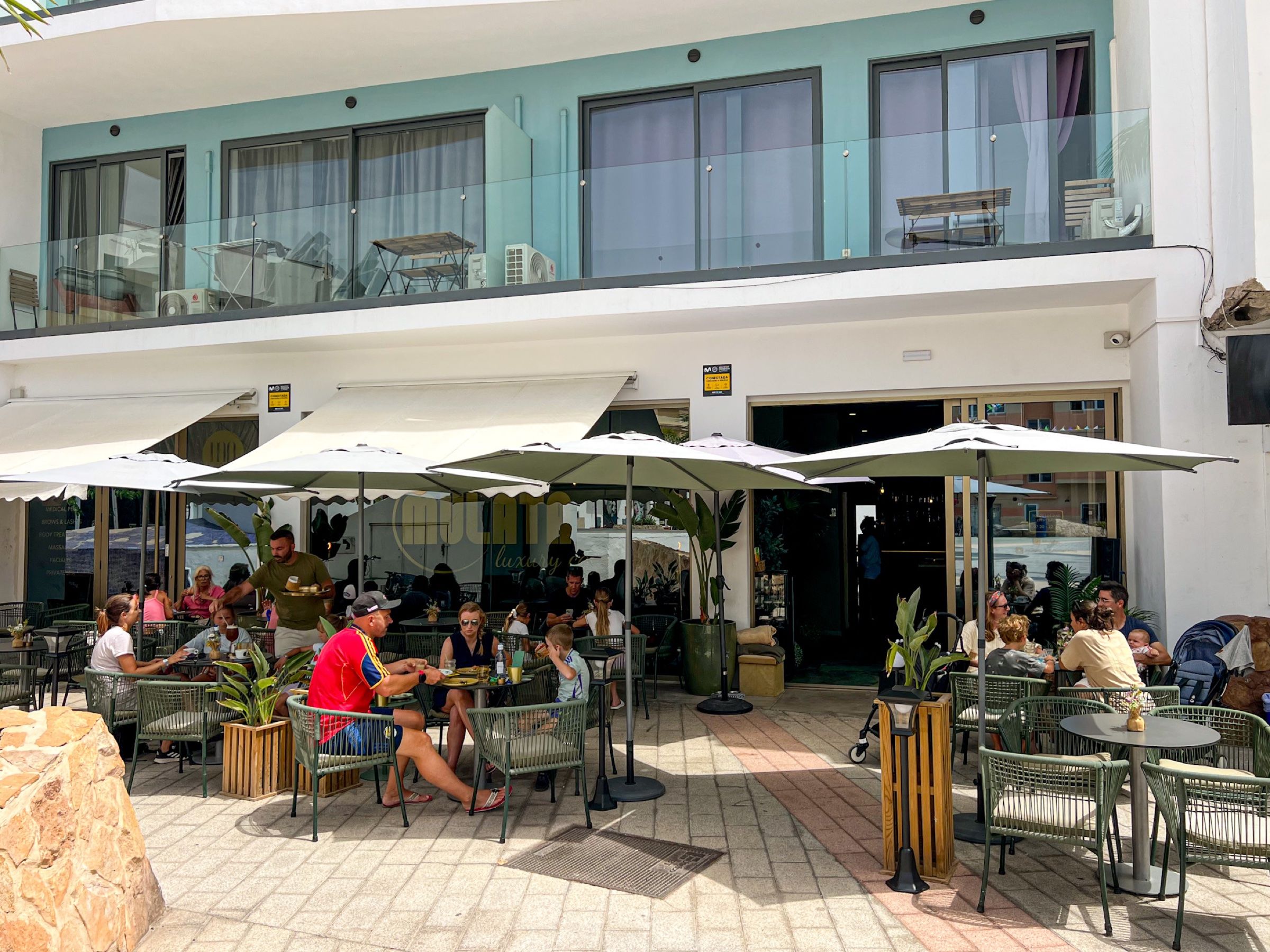Terraza de MULATA Luxury Café en Corralejo (Fuerteventura), con mesas verdes bajo sombrillas blancas, clientes desayunando y la fachada del local al fondo.