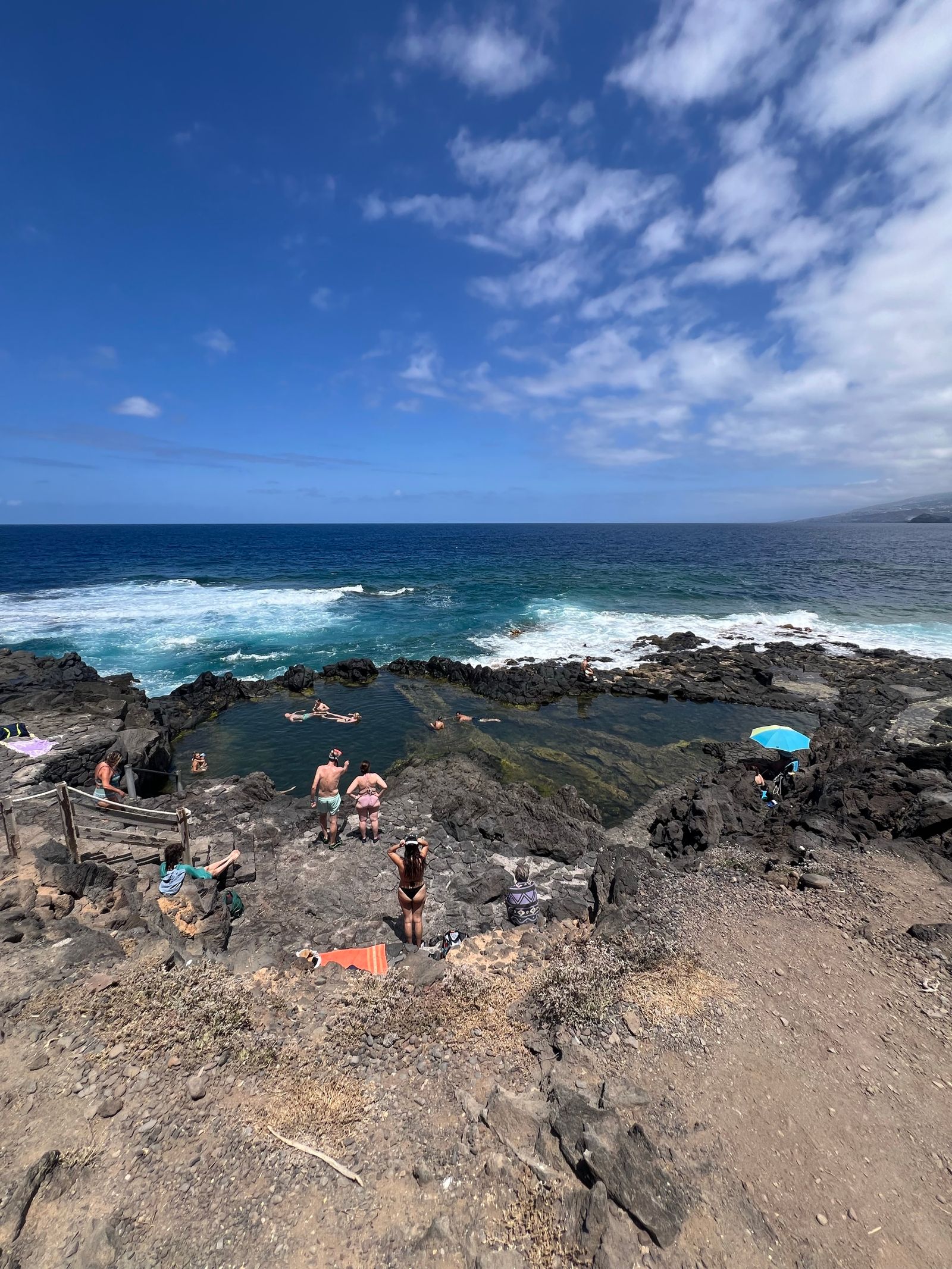 Bañistas flotando en el agua tranquila del Charco de los Chochos, con las olas rompiendo en la roca volcánica detrás.