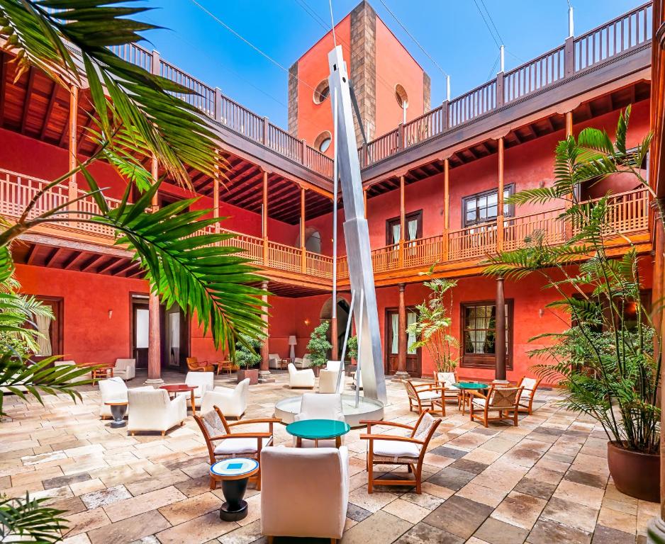 Patio central del Hotel San Roque, en Garachico: paredes rojas, balcones corridos de madera, palmeras en macetas, butacas blancas y la escultura La Centinela en el centro.