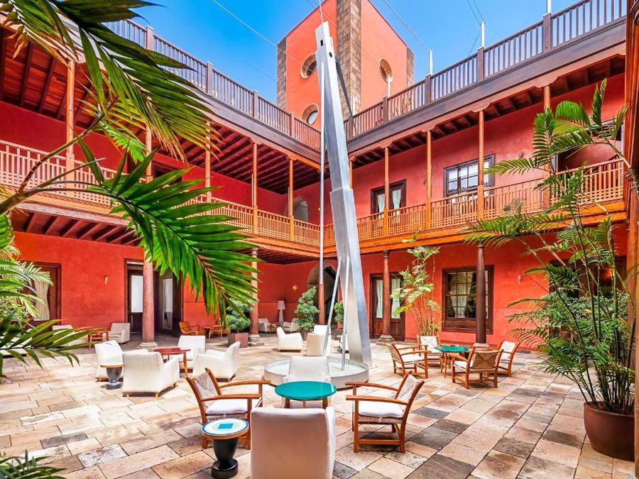 Patio central del Hotel San Roque, en Garachico: paredes rojas, balcones corridos de madera, palmeras en macetas, butacas blancas y la escultura La Centinela en el centro.