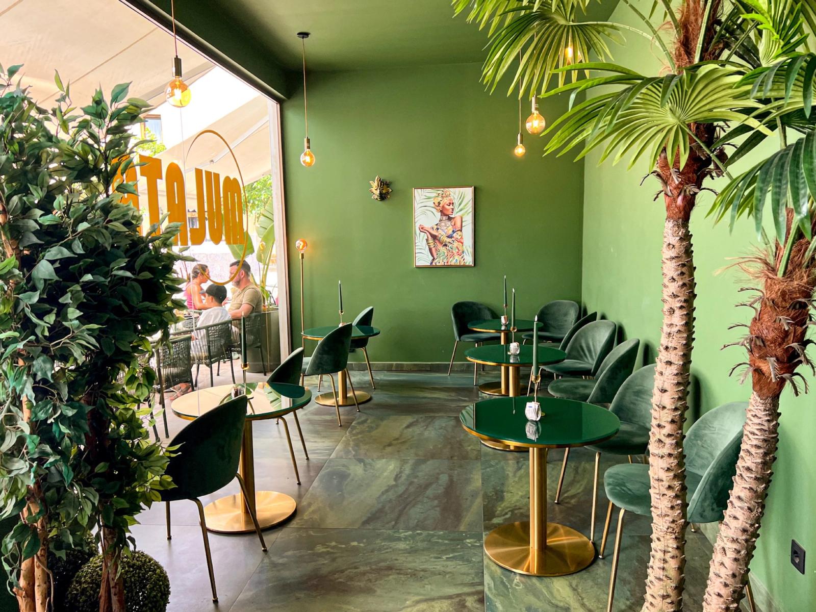 The interior of MULATA Luxury Café in Corralejo: green walls, round tables with green tops and gold bases, velvet chairs, palms and hanging bulbs.