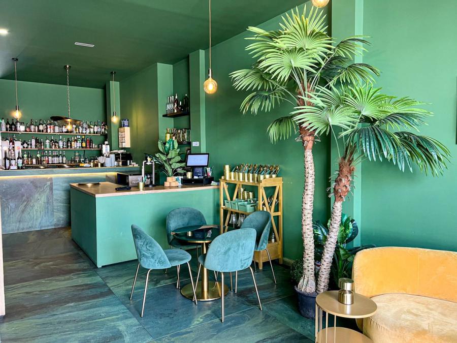 Inside MULATA Luxury Café: the bar with its bottles, green walls, velvet chairs and palms by the window.