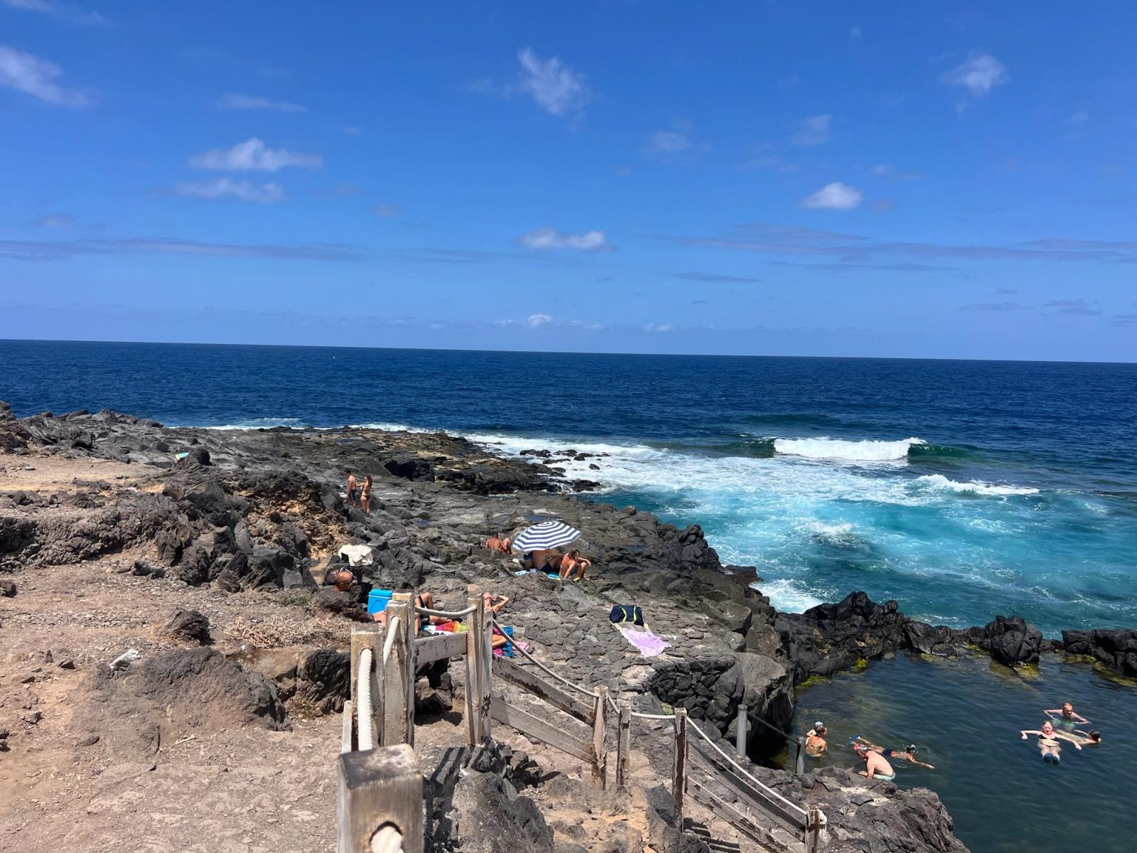Escaleras de madera con barandilla que bajan al Charco de los Chochos, en Los Silos, con gente tomando el sol sobre la roca volcánica y el mar rompiendo detrás.