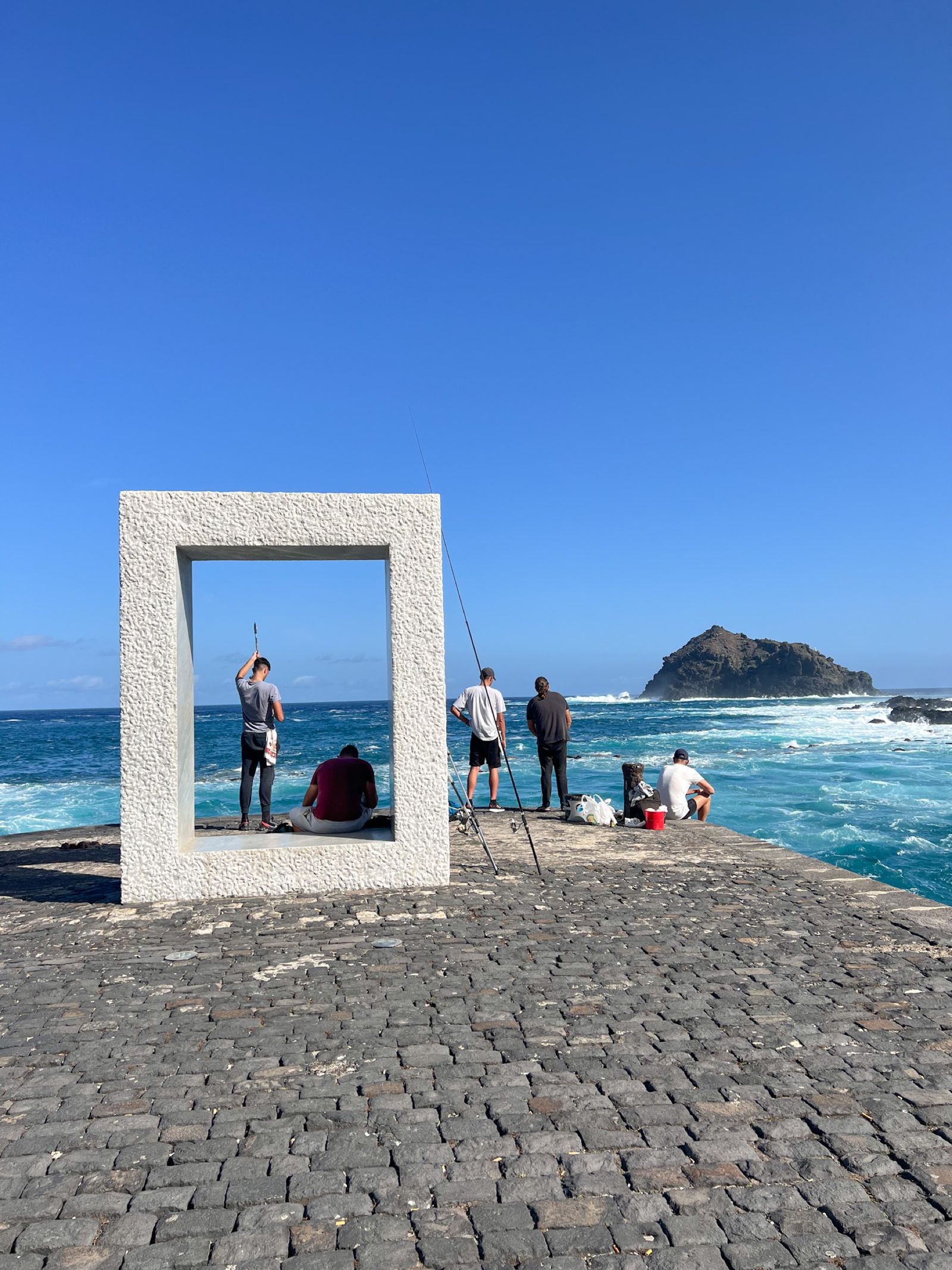 La escultura Tensei Tenmoku de Kan Yasuda en el espigón del muelle de Garachico: un marco de mármol blanco frente al mar, con pescadores echando la caña y el Roque al fondo.