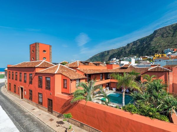 Vista del Hotel San Roque en Garachico: la casona roja con tejados de teja y su piscina rodeada de palmeras, con el mar a un lado y la montaña del pueblo al fondo.