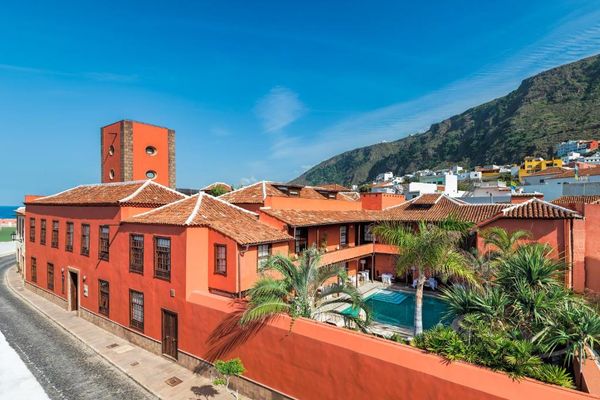 Vista del Hotel San Roque en Garachico: la casona roja con tejados de teja y su piscina rodeada de palmeras, con el mar a un lado y la montaña del pueblo al fondo.