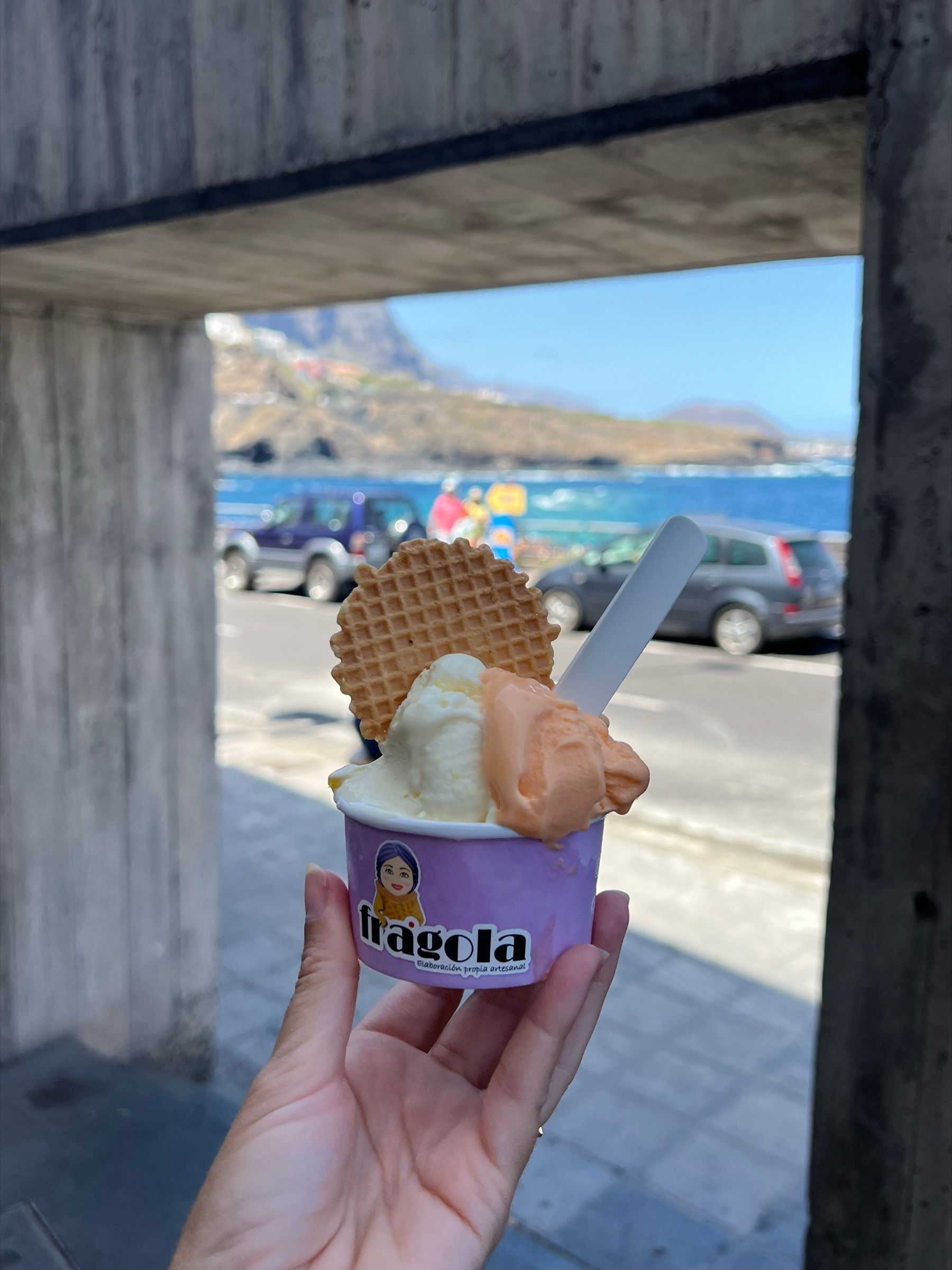 Una tarrina de helado de la heladería Fragola de Garachico, con barquillo, sostenida en la mano frente al mar.