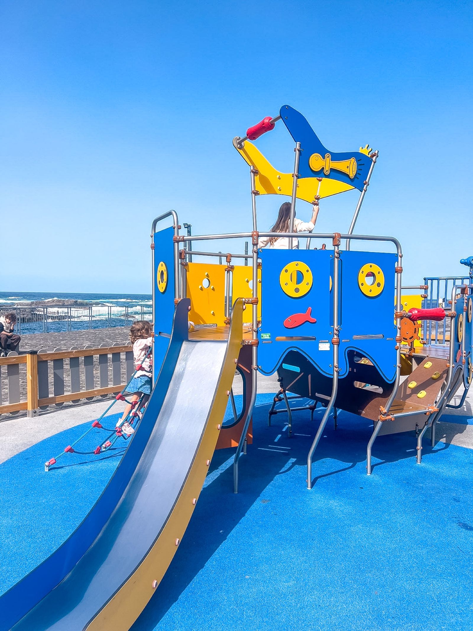 Parque infantil del muelle viejo de Garachico: un barco pirata de madera y metal con tobogán sobre suelo azul, junto a la playa y el mar.