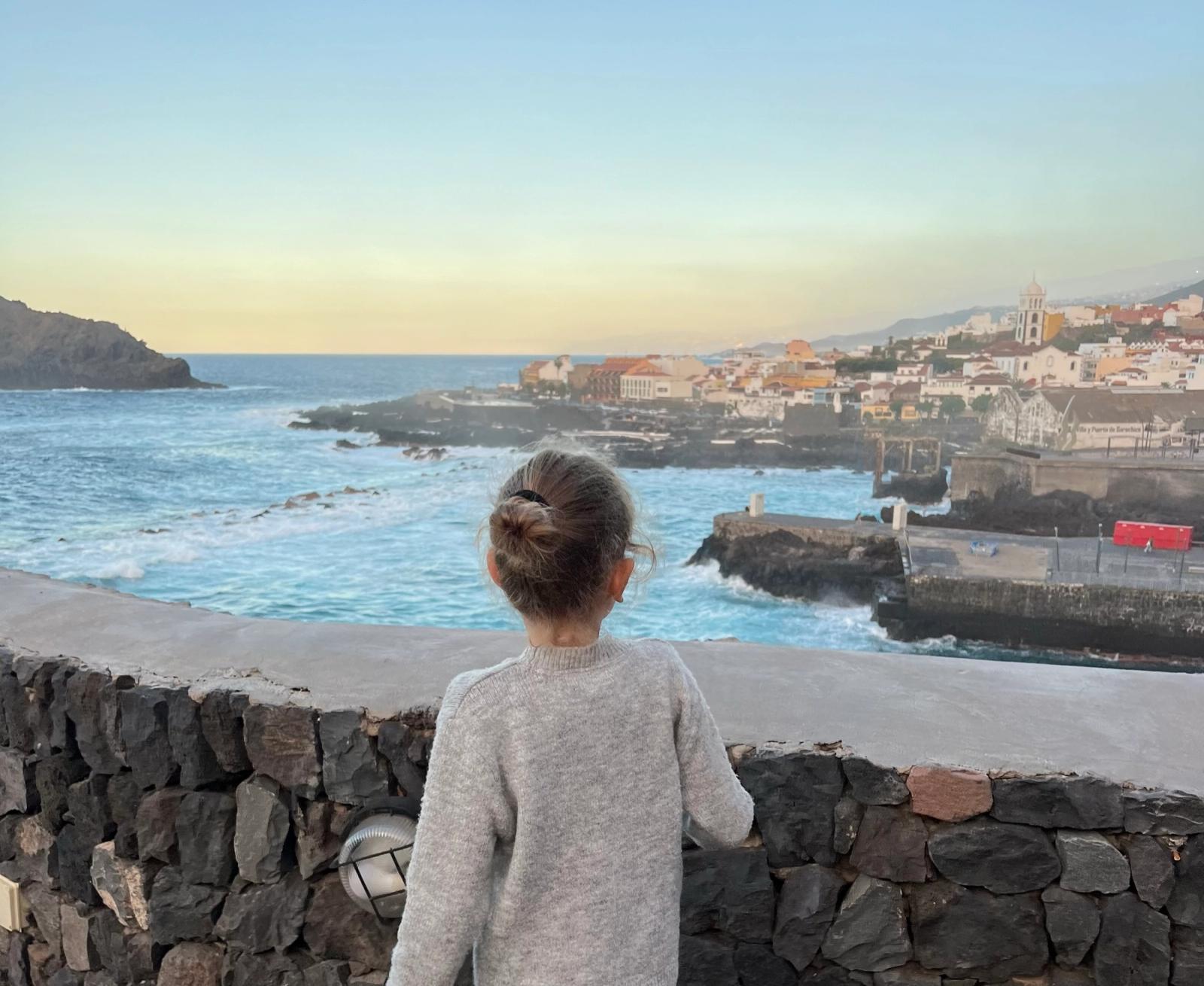 Una niña apoyada en un muro de piedra volcánica mira el puerto y el pueblo de Garachico al atardecer.