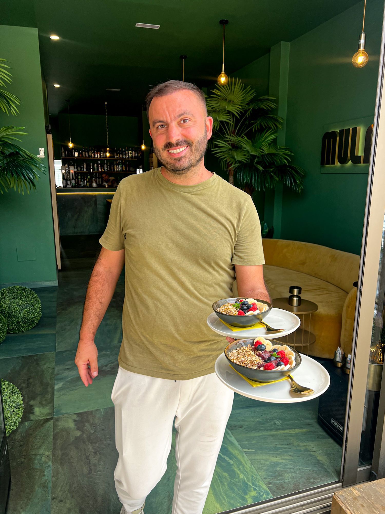 Dragan, the owner of MULATA Luxury Café, in the doorway holding two bowls of fruit and granola.