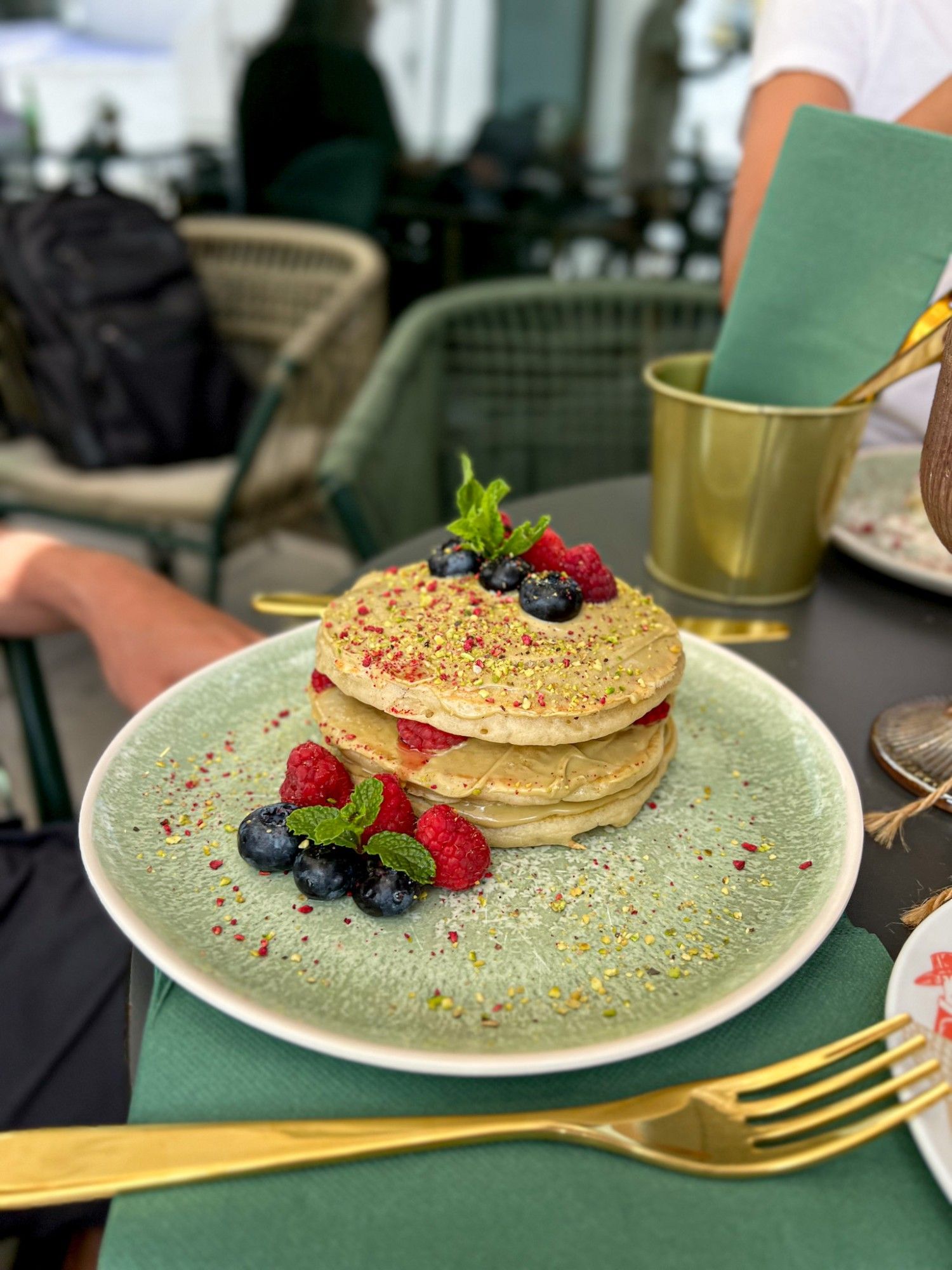 Pistachio pancakes with cream, raspberries, blueberries and mint, sprinkled with chopped pistachio, on a green plate on the terrace table.