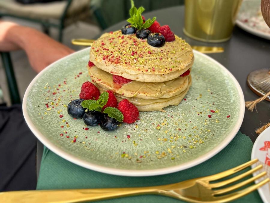 Pistachio pancakes with cream, raspberries, blueberries and mint, sprinkled with chopped pistachio, on a green plate on the terrace table.