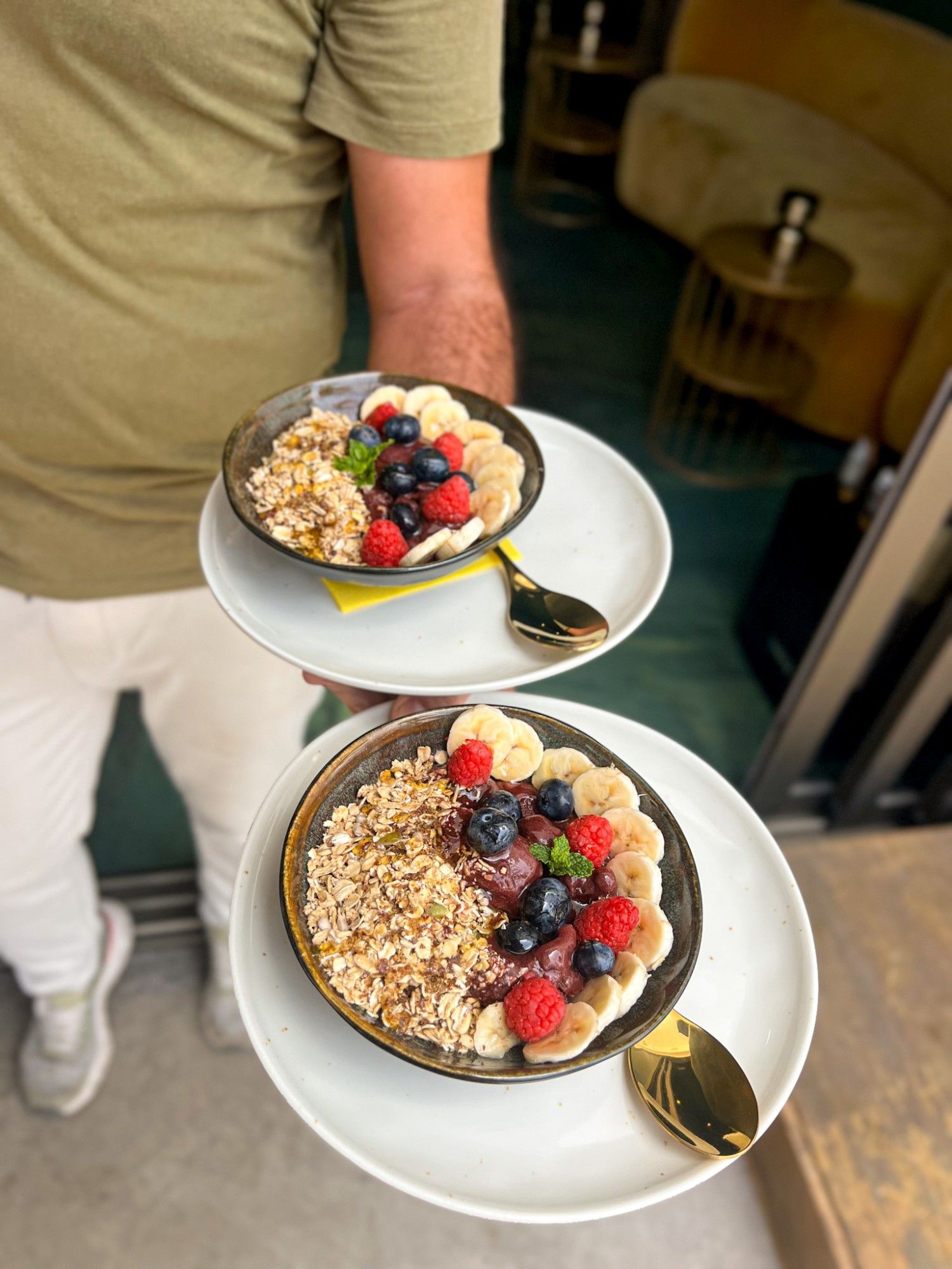 Two bowls with a red fruit base, granola, banana, raspberries and blueberries, served in dark bowls with gold spoons.