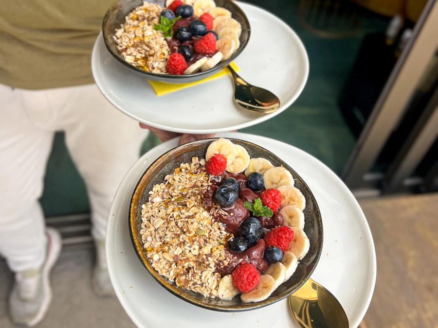 Two bowls with a red fruit base, granola, banana, raspberries and blueberries, served in dark bowls with gold spoons.