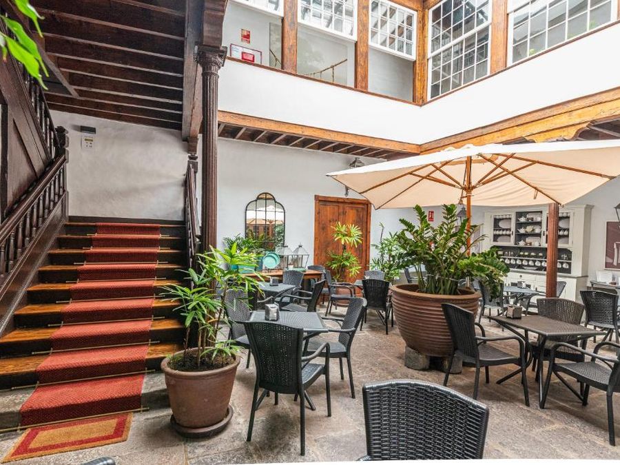 Patio interior de Isla Baja Suites, en Garachico: escalera de madera con alfombra roja, mesas bajo una sombrilla y plantas en macetas, en una casa canaria del siglo XVIII.