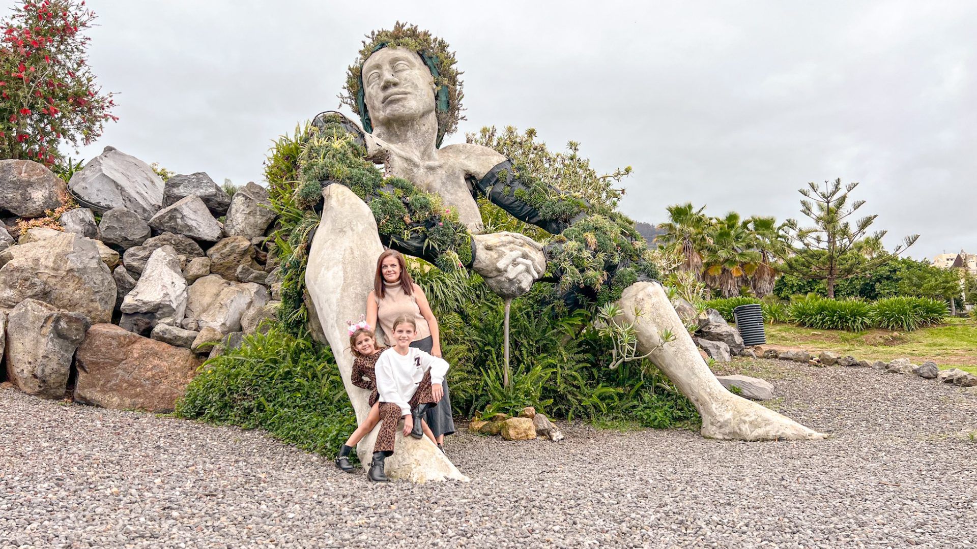 La escultura de La Giganta de Santa Úrsula, recostada sobre las rocas y cubierta de vegetación, con Miriam y las niñas posando a sus pies.
