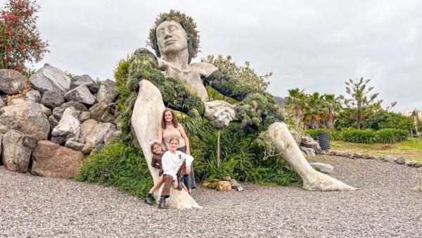 La escultura de La Giganta de Santa Úrsula, recostada sobre las rocas y cubierta de vegetación, con Miriam y las niñas posando a sus pies.