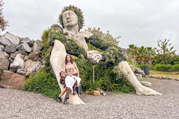 La escultura de La Giganta de Santa Úrsula, recostada sobre las rocas y cubierta de vegetación, con Miriam y las niñas posando a sus pies.