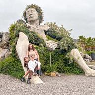 La escultura de La Giganta de Santa Úrsula, recostada sobre las rocas y cubierta de vegetación, con Miriam y las niñas posando a sus pies.
