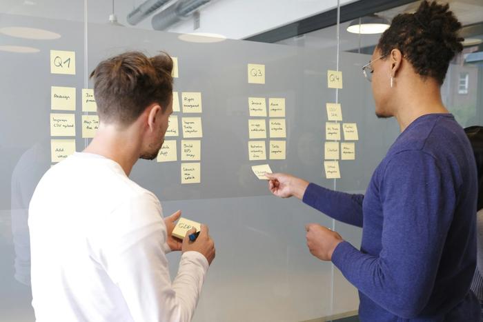 two men are standing in front of a glass wall with sticky notes on it .