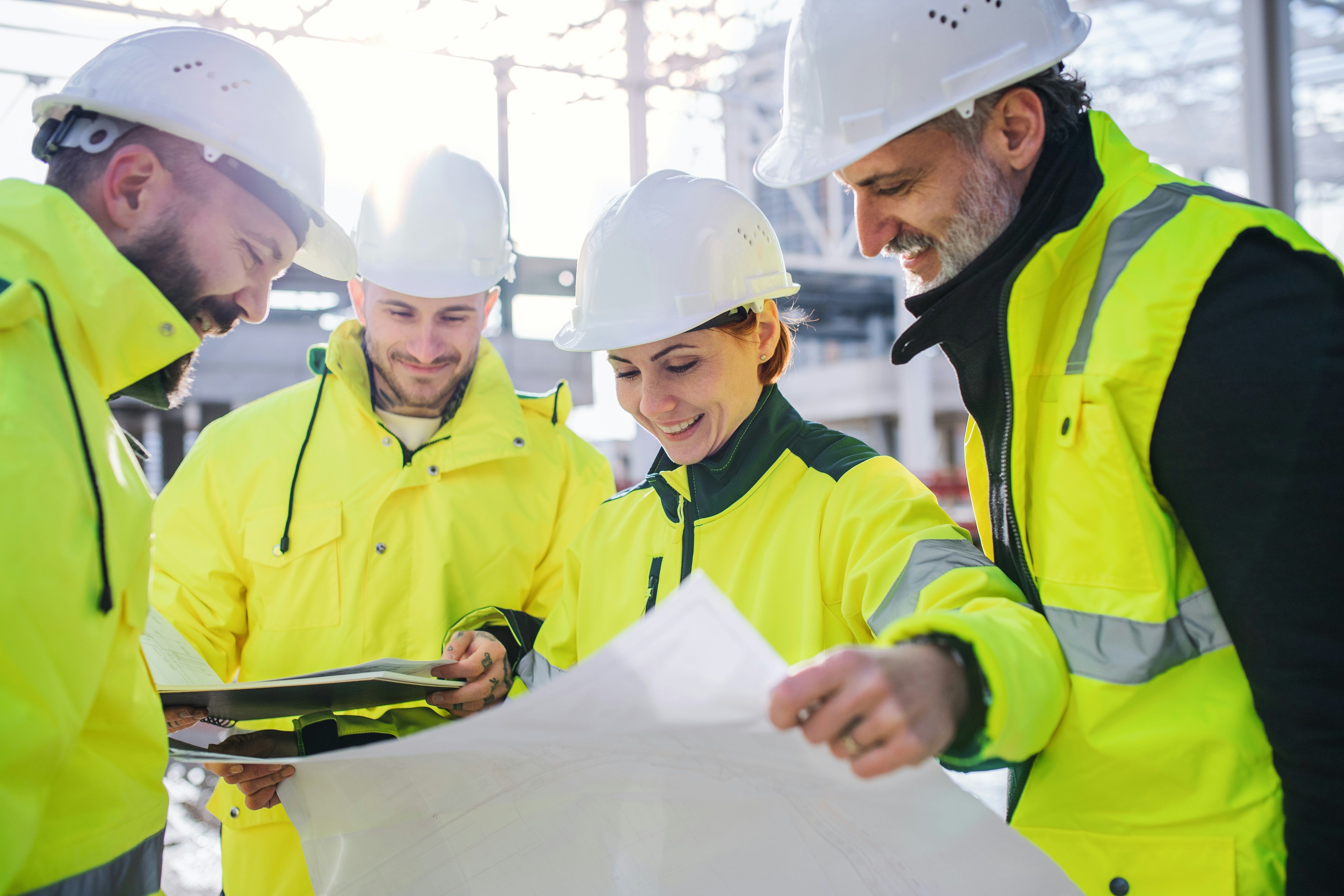 Quatro operários da construção civil, usando capacetes e coletes de alta visibilidade, analisam plantas arquitetônicas.