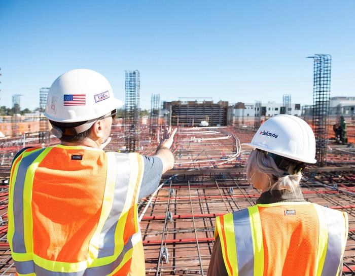 Two people in hard hats and safety vests on a construction site, one pointing.