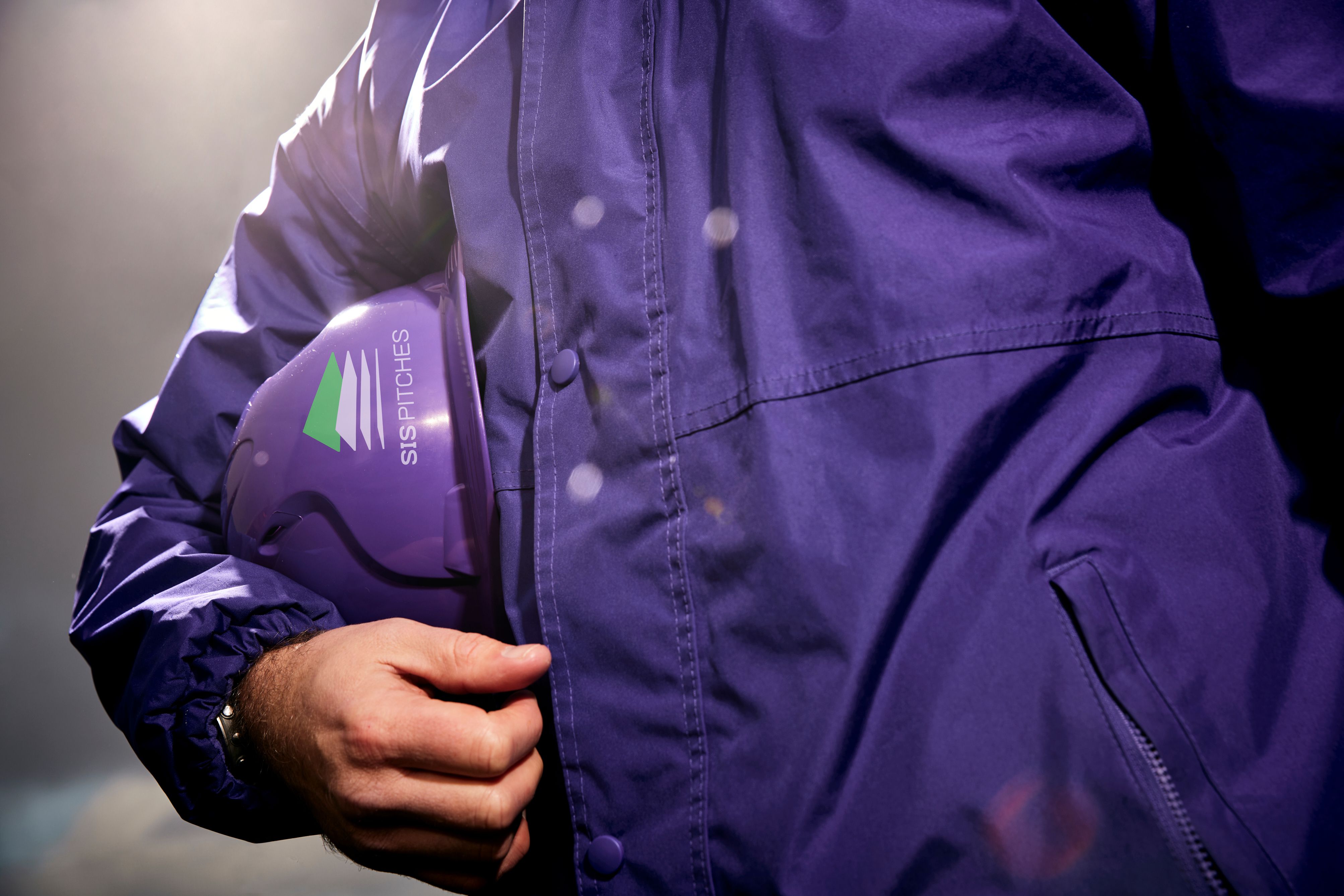 Person in a purple jacket holding a purple hard hat with a SIS PITCHES logo.