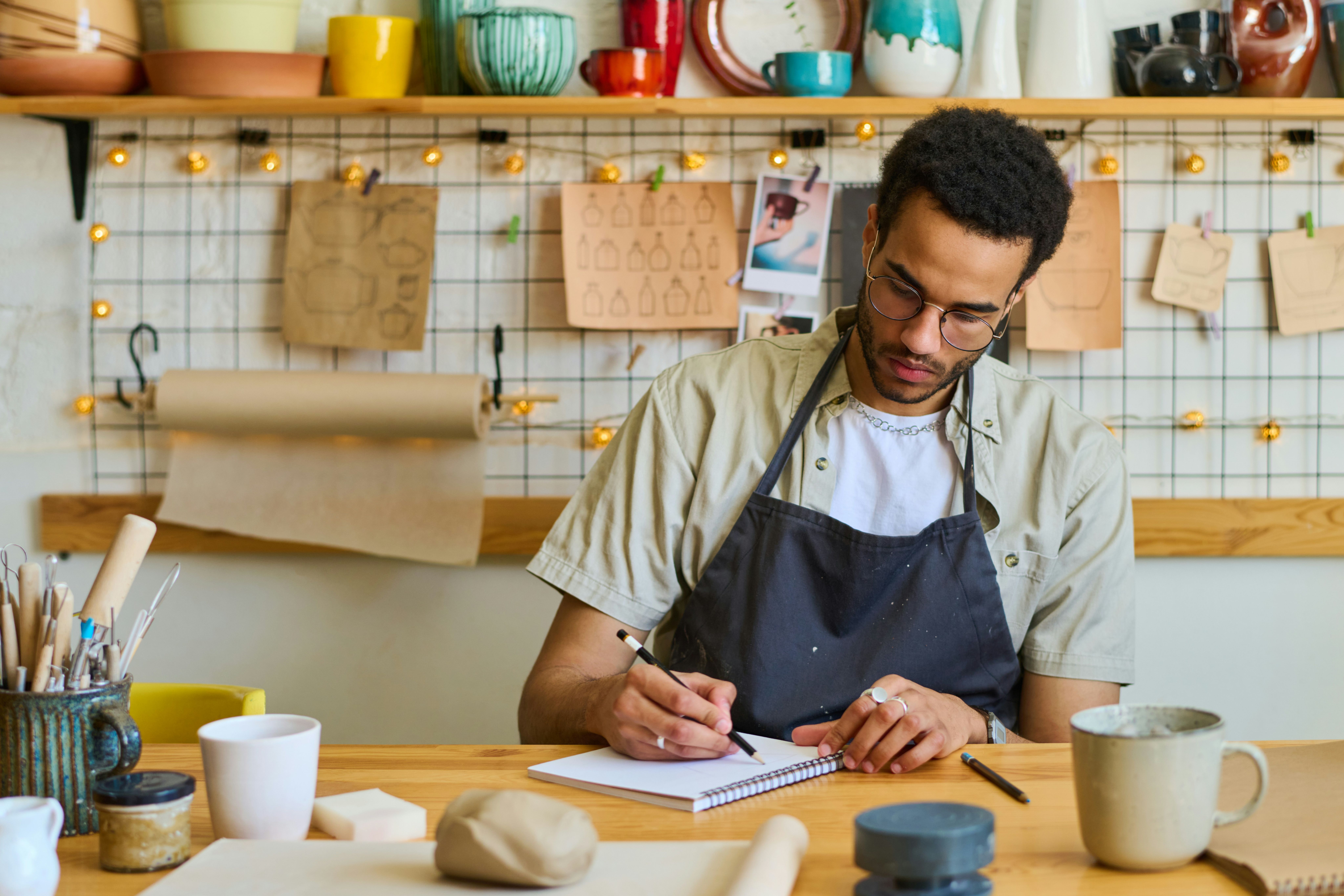 a man making writing in a notebook while sat at a desk surrounded by pottery crafting tools