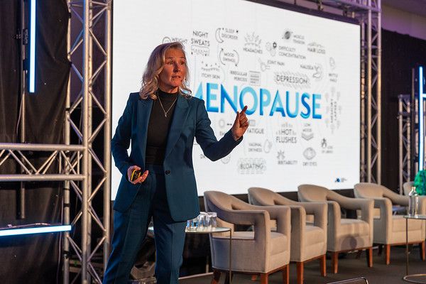Lynda presenta en el escenario frente a una gran pantalla que muestra "MENOPAUSIA" y sus síntomas.