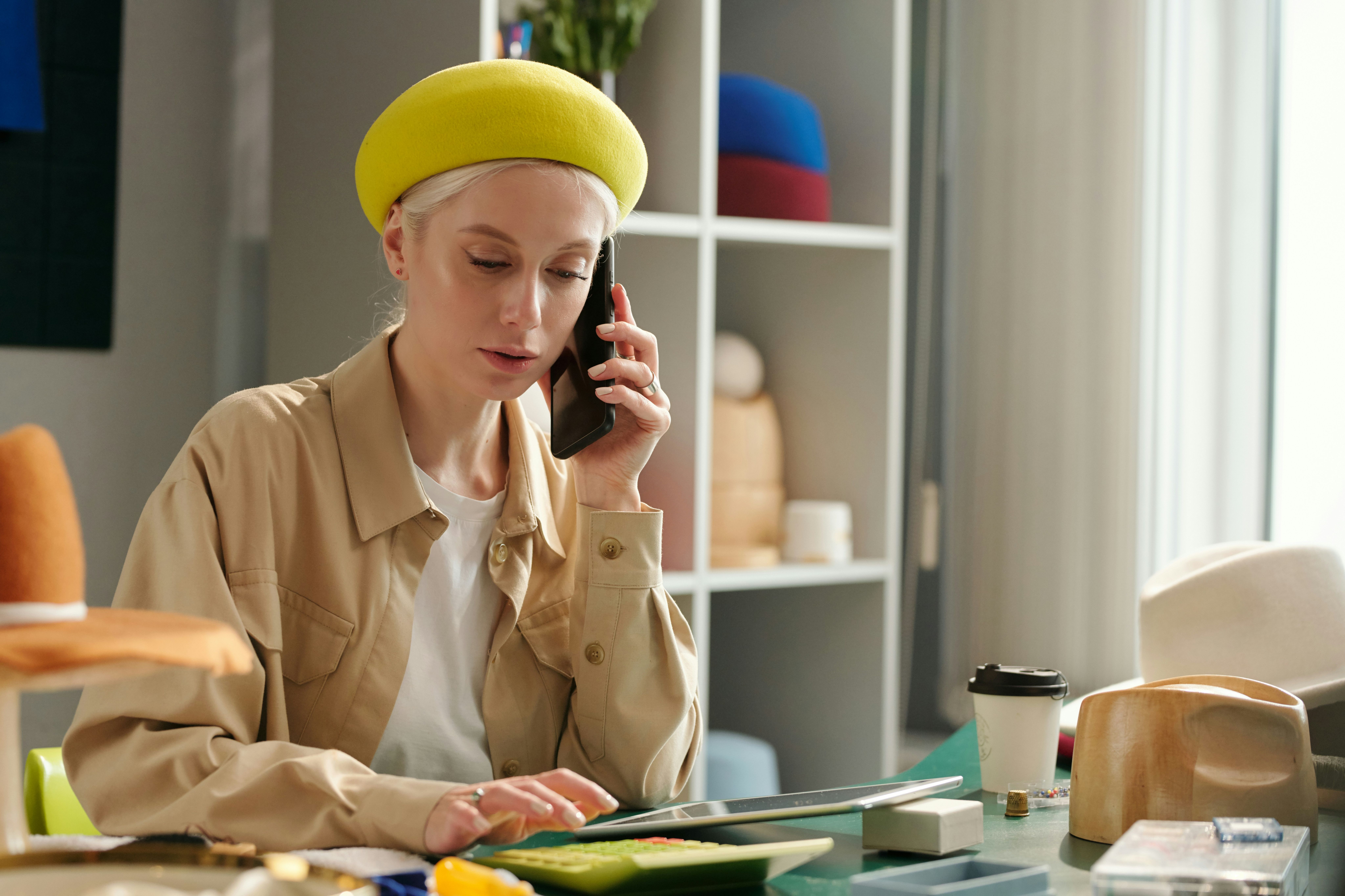 a woman in a yellow beret is sitting at a desk talking on a cell phone .