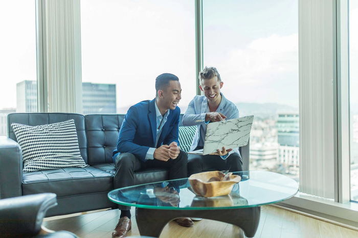 two men sit on a couch looking at a laptop
