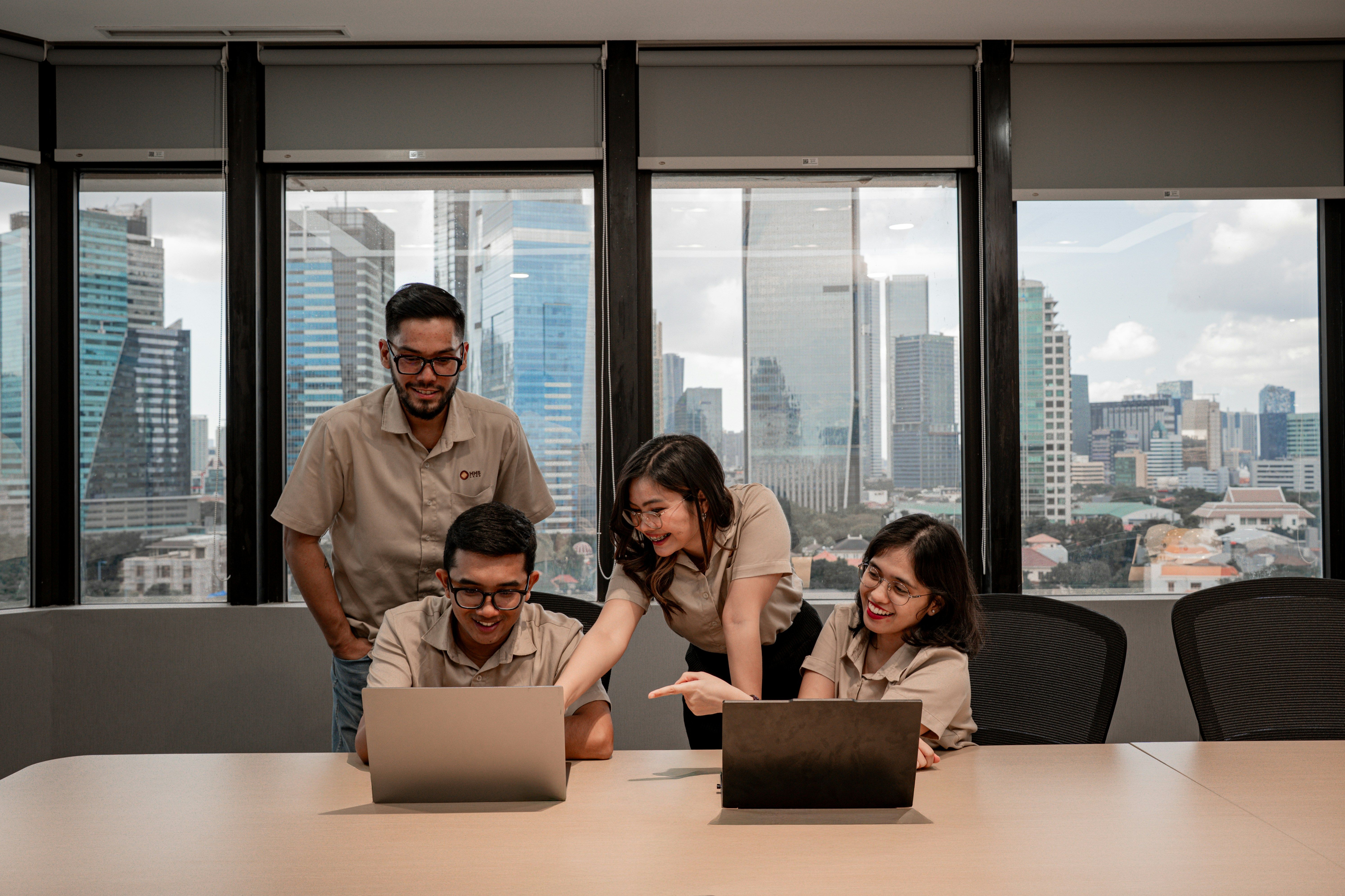 Quatro colegas colaborando em laptops em um escritório com vista para o horizonte da cidade.