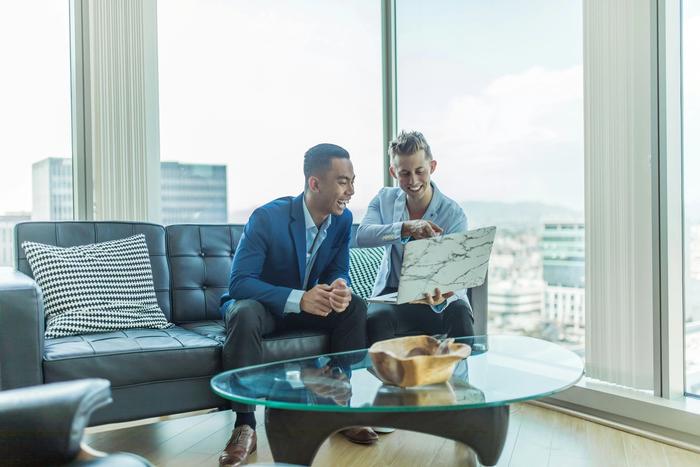 two men are sitting on a couch looking at a laptop .