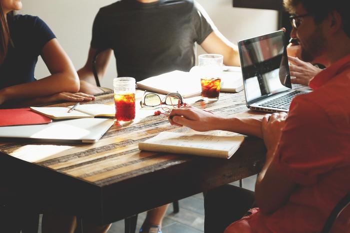 A group of people are sitting around a wooden table with laptops and notebooks.